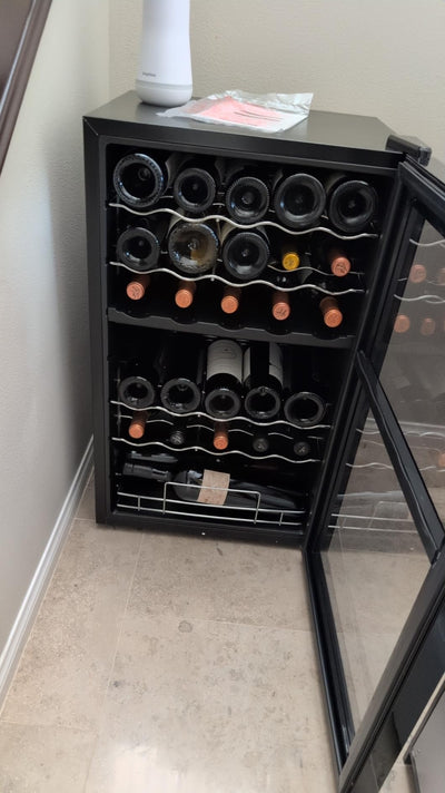 Black wine cooler with open door displaying wine bottles on a light-colored floor.